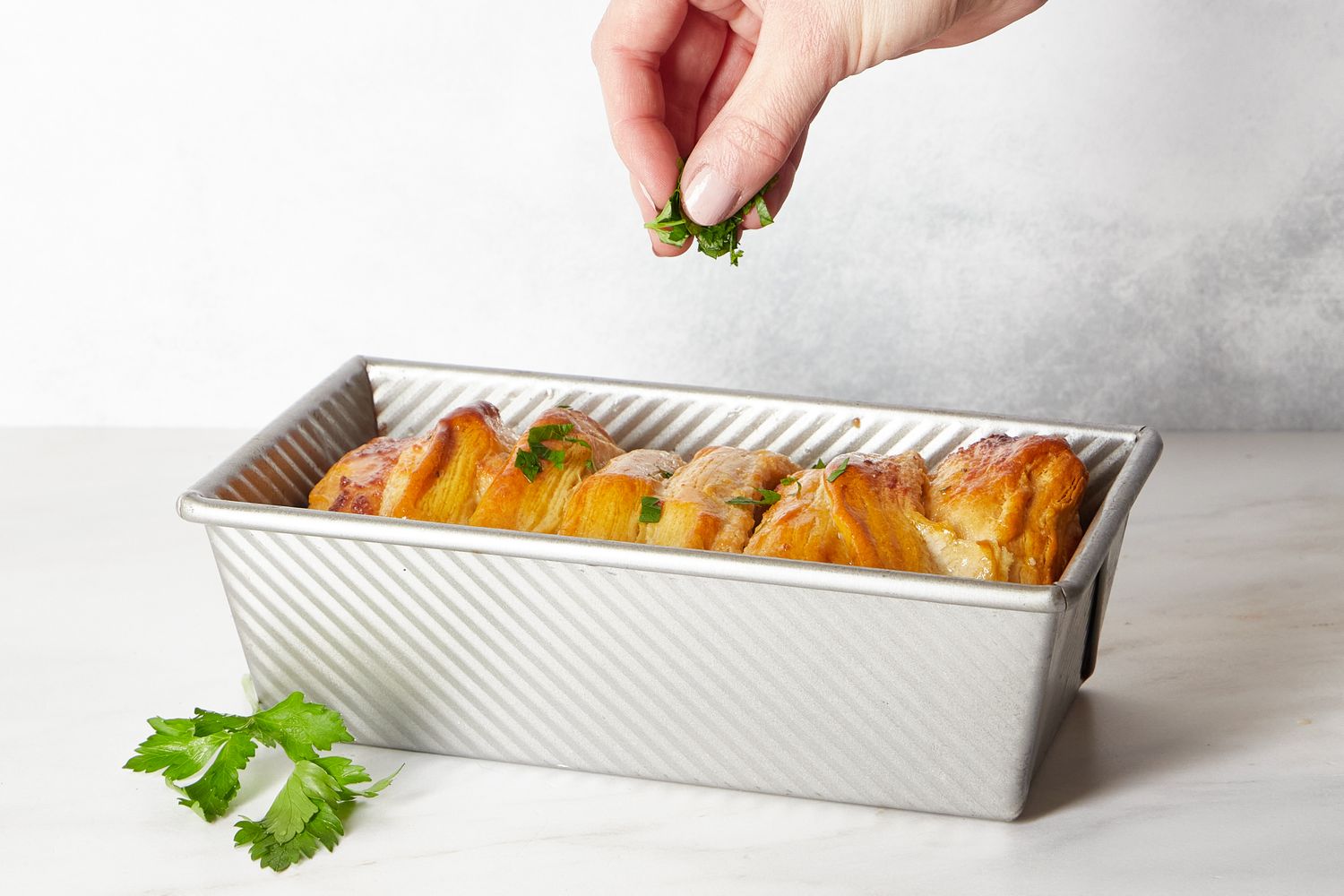 A hand sprinkling herbs on pullapart bread in a rectangular baking tin