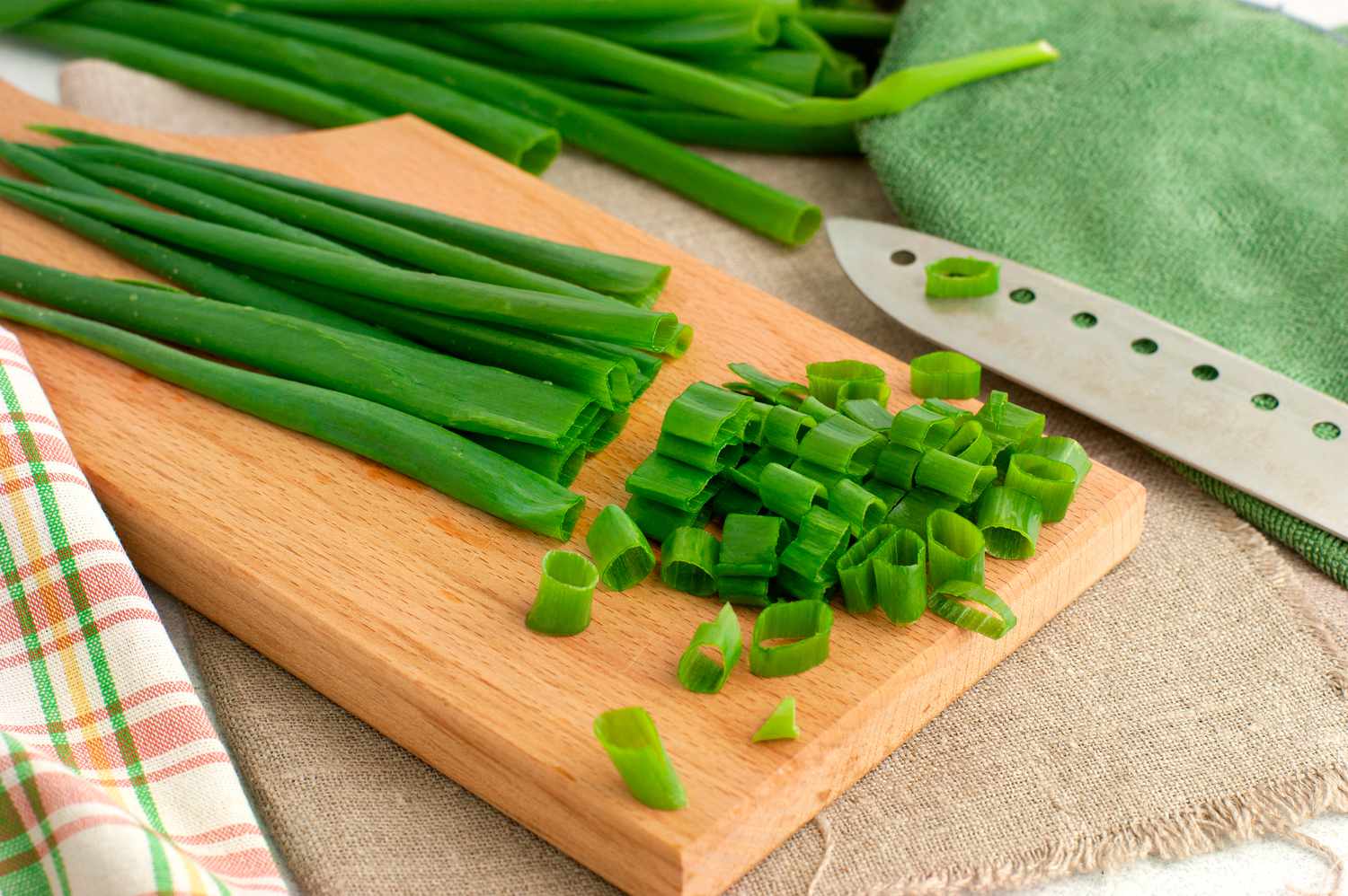 Chopped green onions on a wooden cutting board next to a knife and resting on green, tan and plaid cloth napkins