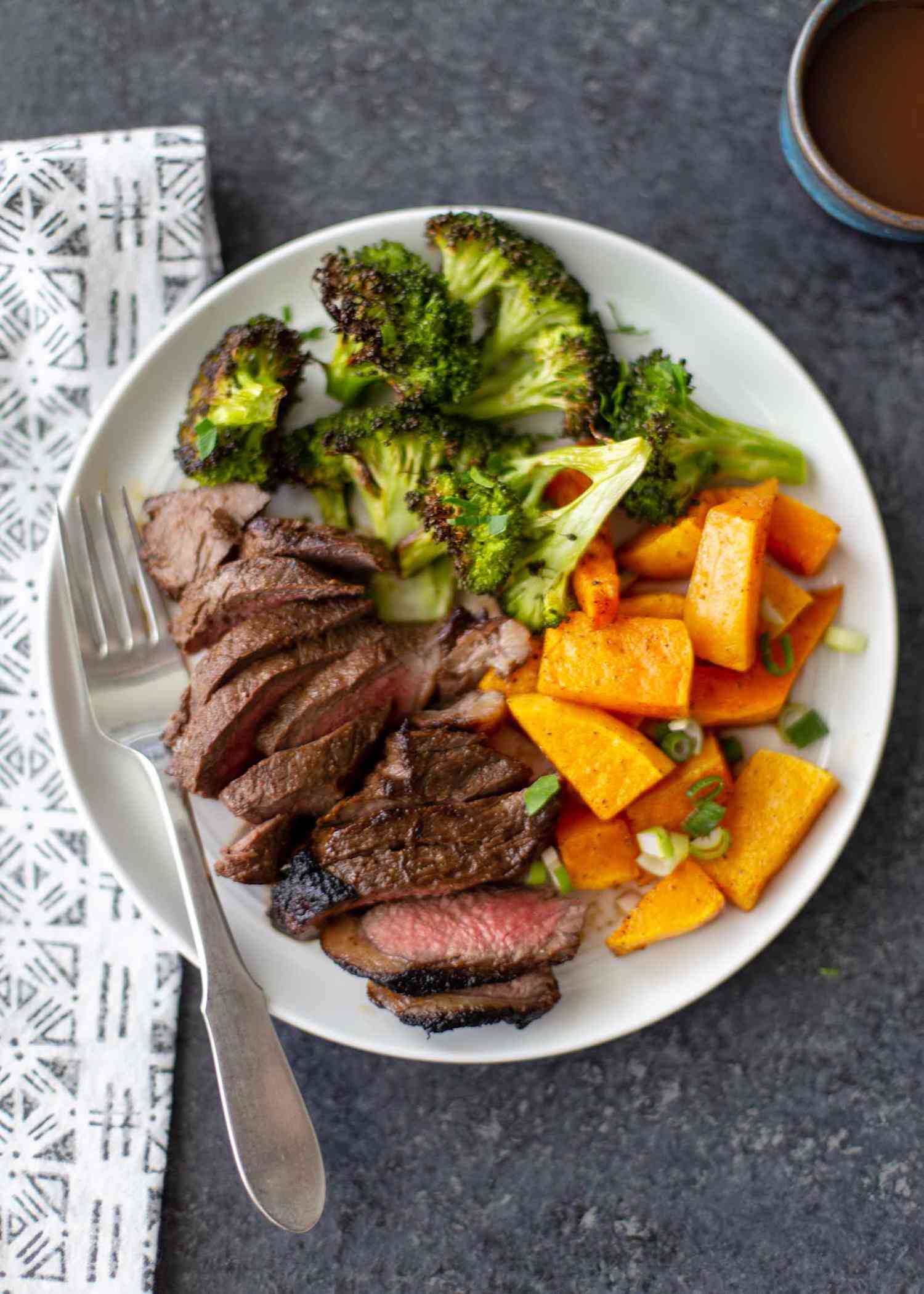 Sliced steak cooked in the stove on a white plate with seared edges and pink middle. Roasted broccoli and squash are on the plate.