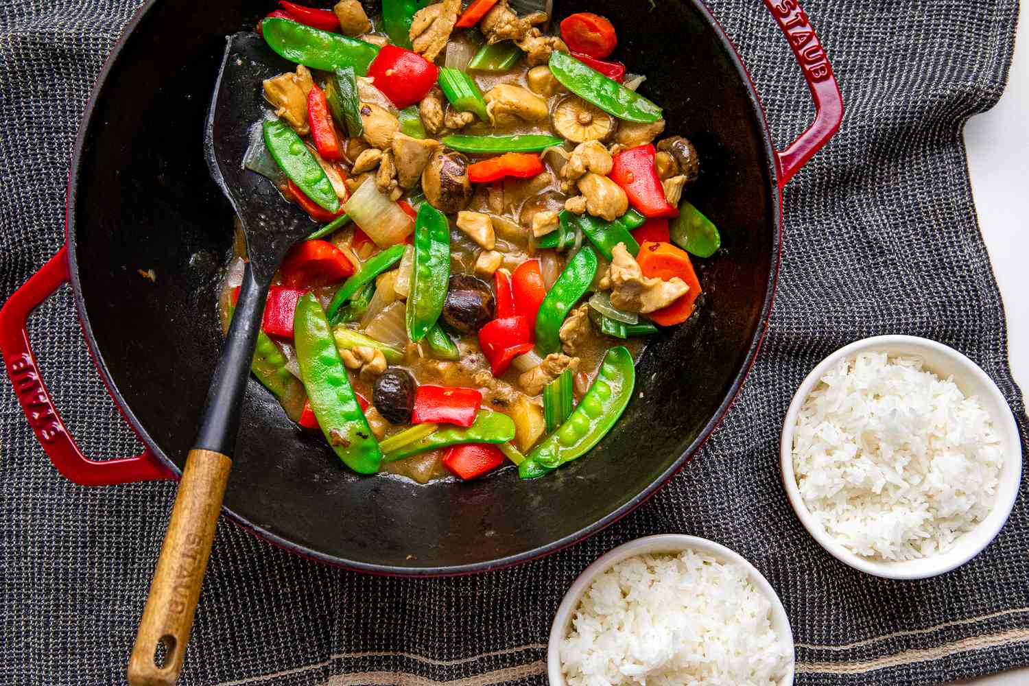 Chop Suey in a Staub Wok With a Spatula, and Next to It, Two Bowls of Rice on a Grey Kitchen Towel