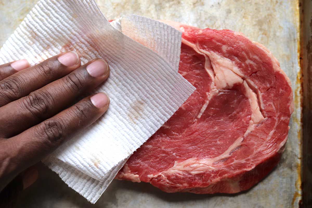 Raw steak on a worn baking sheet being patted dry with a paper towel to show how to cook steak on the stovetop.