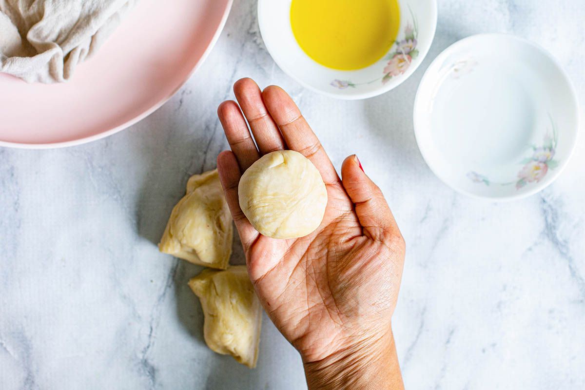 A ball of dough in a woman's hand with a bowl of butter nearby and a pink plate.