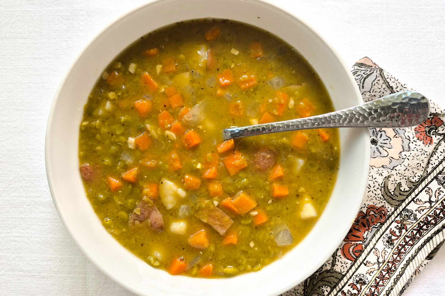 A bowl of split pea soup with diced vegetables and a spoon on a table next to a patterned napkin