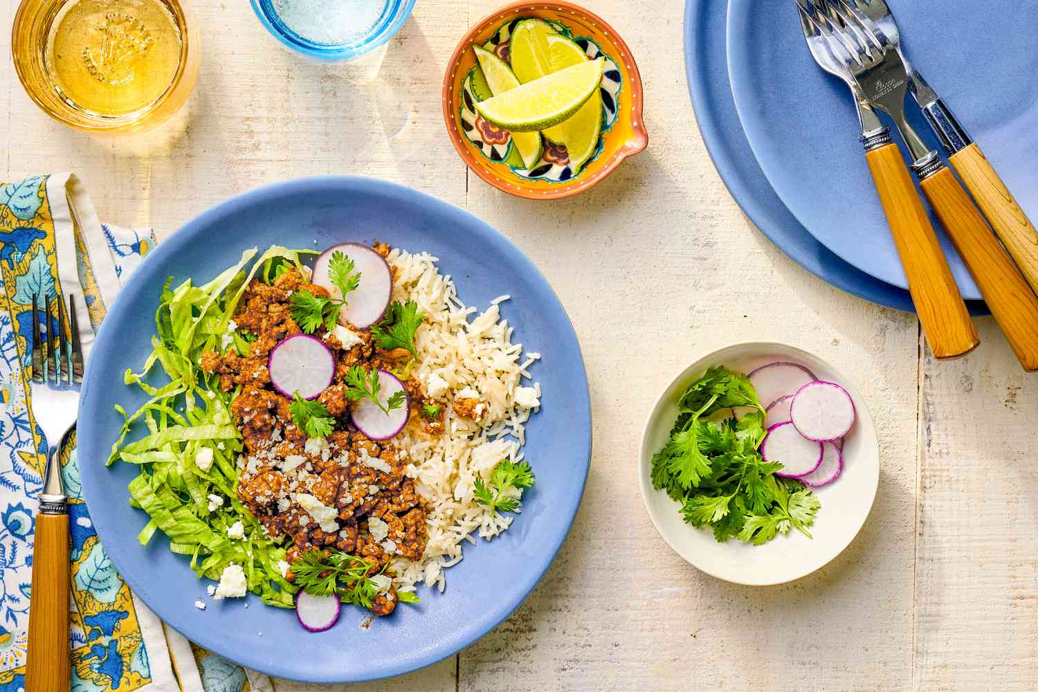 Plate of 5-ingredient turkey taco bowl at a table setting with glasses of water, bowl of lime wedges, a fork on a table napkin, a bowl of cilantro and sliced radish, and stack of plates and forks