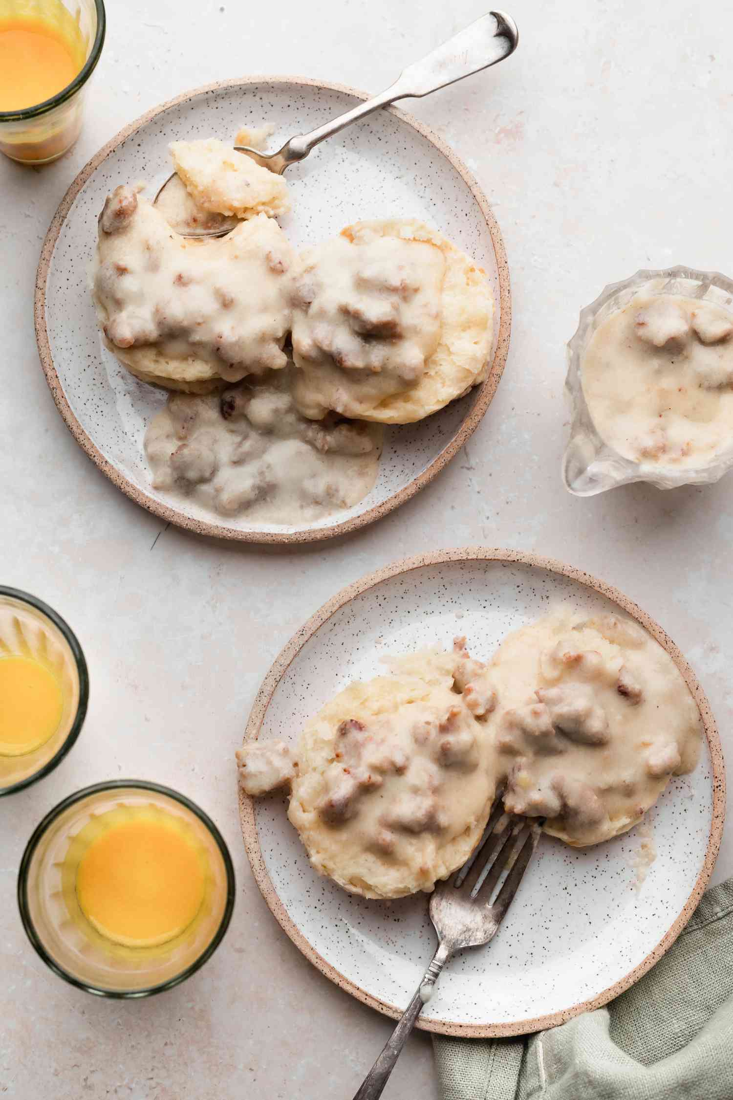 Overhead view of two plates of sausage gravy and biscuits served with orange juice.