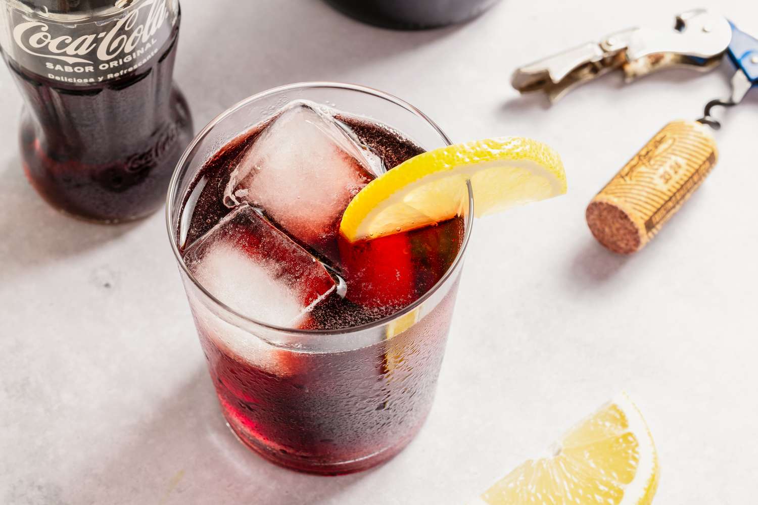 A glass of Kalimotxo with ice cubes and a lemon wedge, a cola bottle and a wine cork on the side