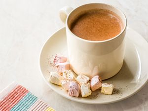 A cup of hot cocoa on a saucer with marshmallows placed next to a striped fabric