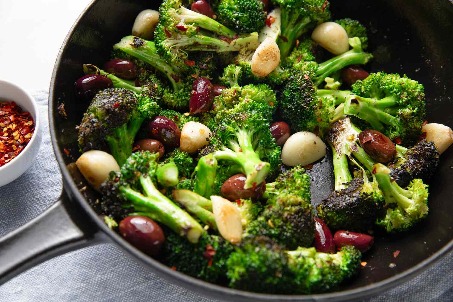 Angled view of a cast iron skillet of seared broccoli with spices, garlic cloves and olives next to a small bowl of spices