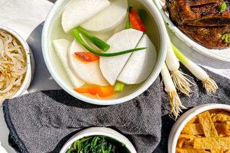 Bowl of Dongchimi Surrounded by Bowls of Banchan (Side Dishes) and a Bowl of Korean Short Ribs. On the Table, a Table Napkin and Some Whole Green Onions.