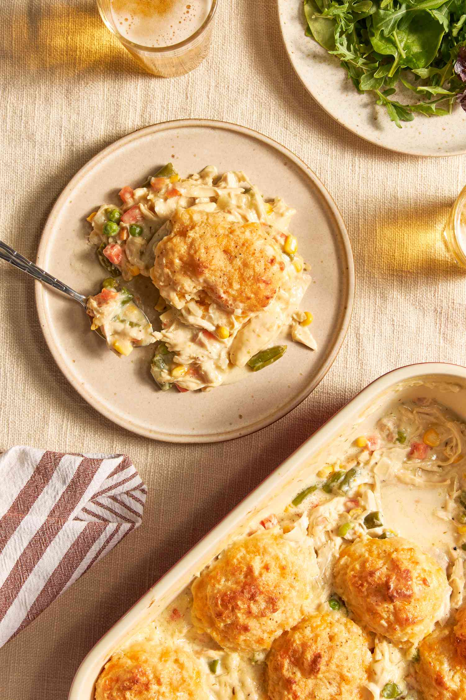 Serving of Chicken Cobbler With Cheddar Bay Biscuit Top on a plate at a table setting with a plate of salad greens, glasses of cider, and a casserole dish with cobbler