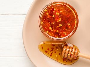 Jar of chili honey with red pepper flakes next to a honey dipper coated with the spiced honey