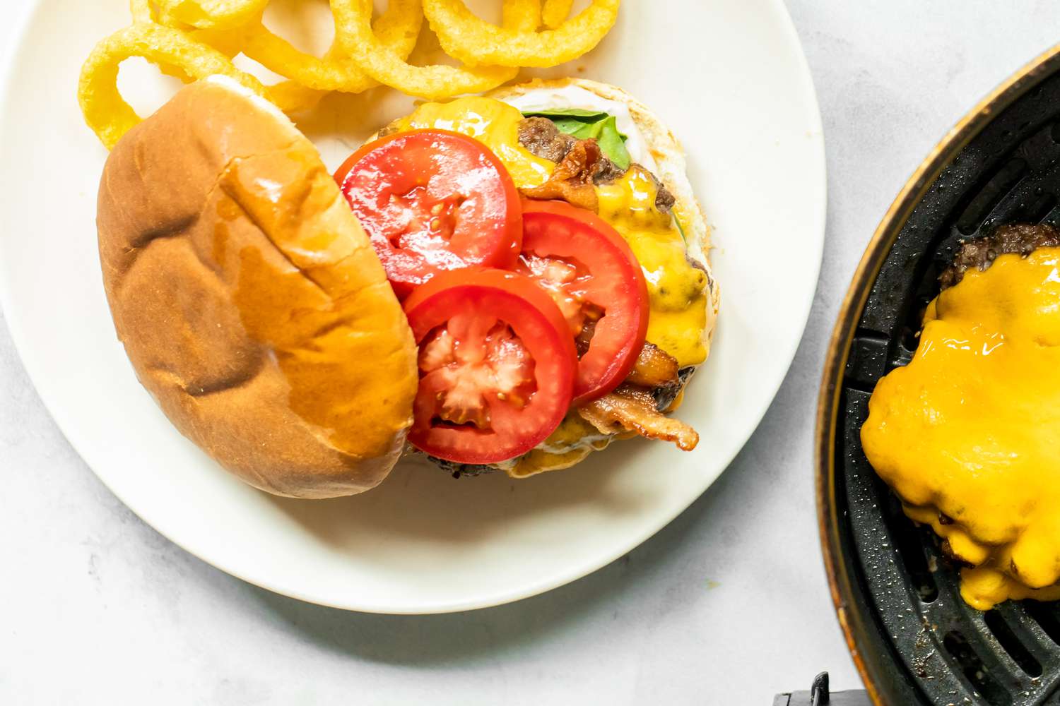Air Fryer Burger with Onion Rings on a Plate Next to Air Fryer Basin with More Burger Patties