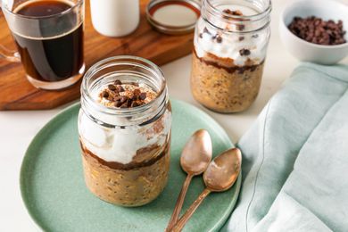 Jar of tiramisu overnight oats topped with Greek yogurt, mini chocolate chips, and cocoa powder on a small plate with two spoons, and in the surroundings, a bowl of mini chocolate chips, another jar of overnight oats, and a wooden board with a glass of coffee, a glass of milk, and the jar lid