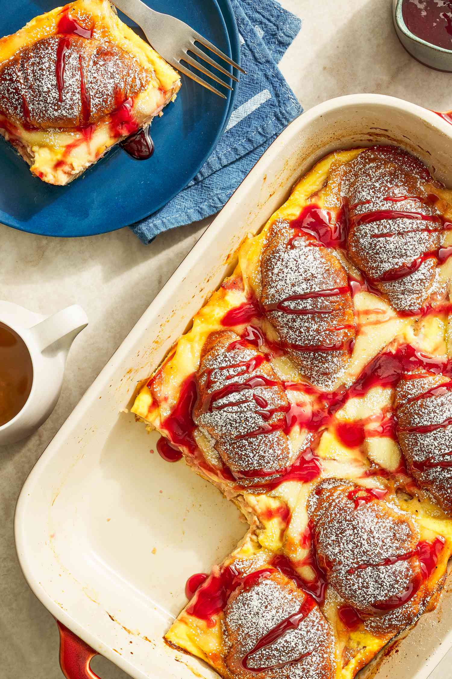 Monte Cristo croissant casserole in a casserole dish and in the surroundings, a serving on a small plate with a fork, a container of jelly, and cups of coffee