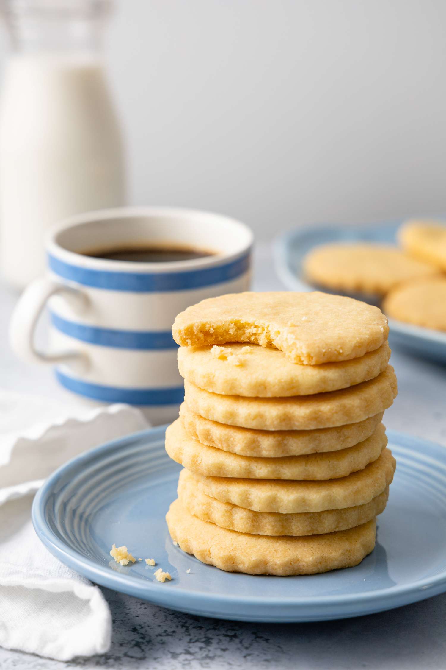 Stack of Classic Shortbread Cookies with the Top Cookie Missing a Bite and a Cup of Coffee in the Background
