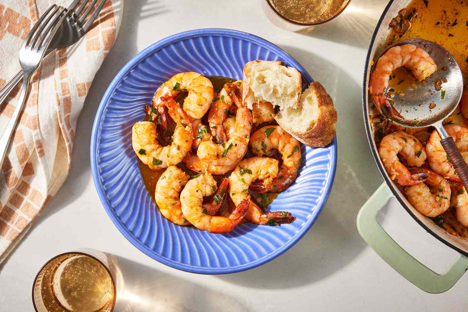 bowl of spanish garlic shrimp with baguette slices at a table setting with forks on a table napkin, glasses of beer, and more shimp in the dutch oven