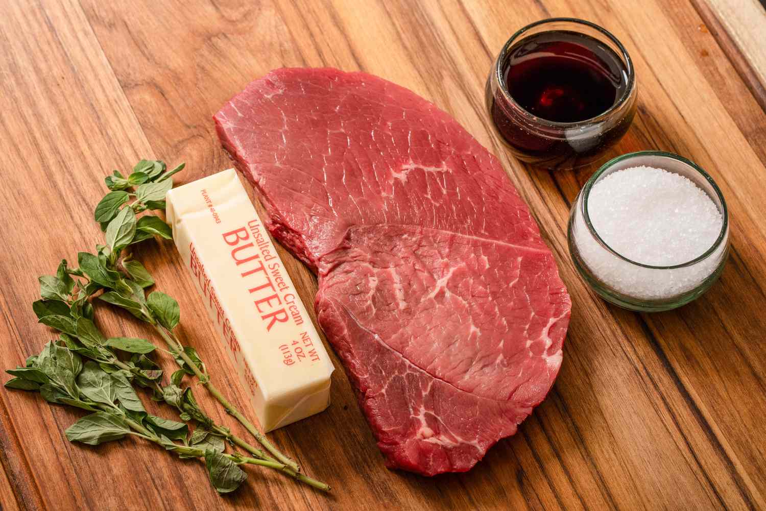 A raw steak, butter, fresh herbs, red wine, and salt arranged on a wooden surface