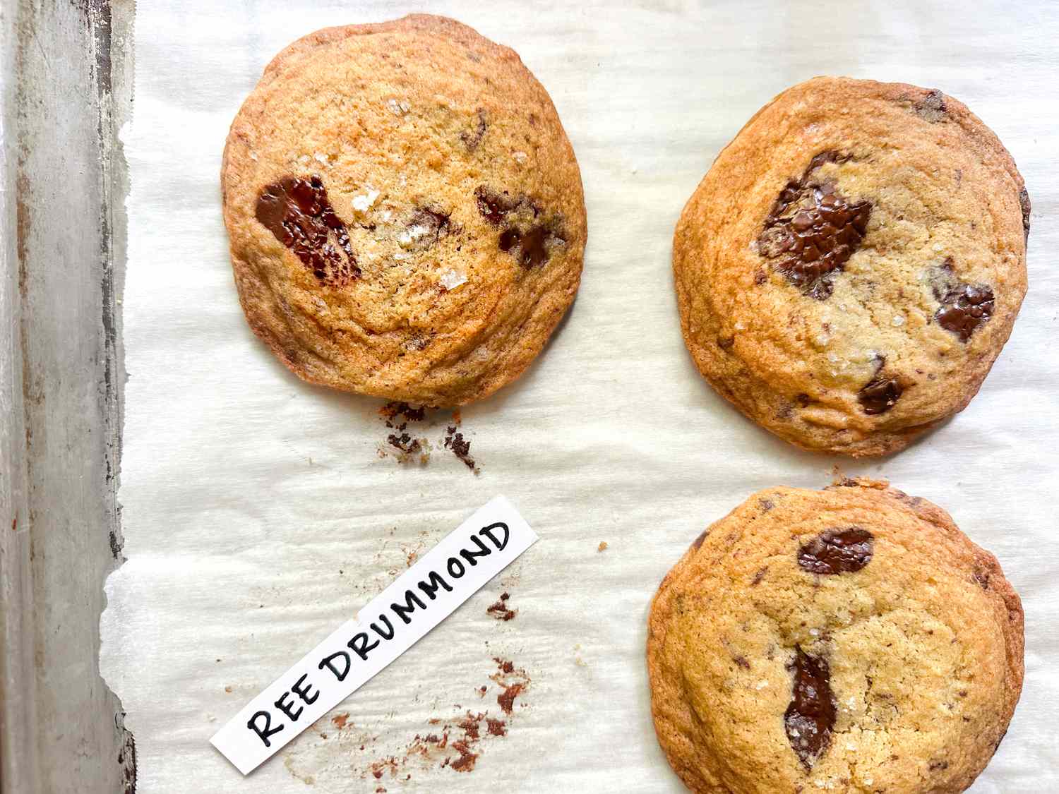 Three baked chocolate chip cookies labeled as Ree Drummonds recipe on a parchmentlined baking tray