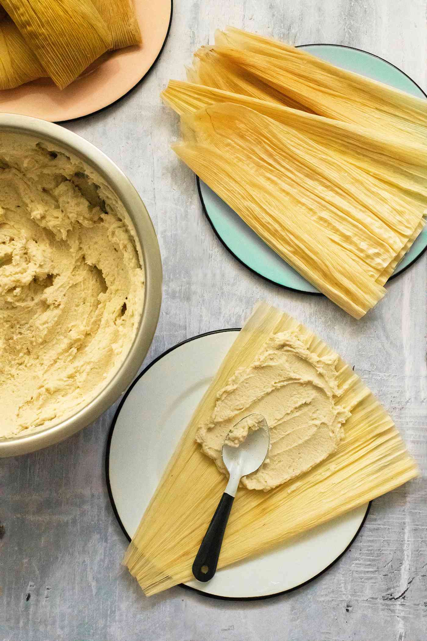 Bowl of Masa (Tamales Dough) Next to a Plate with a Corn Husk Smeared with Masa Using the Back of the Spoon and a Plate with More Corn Husks