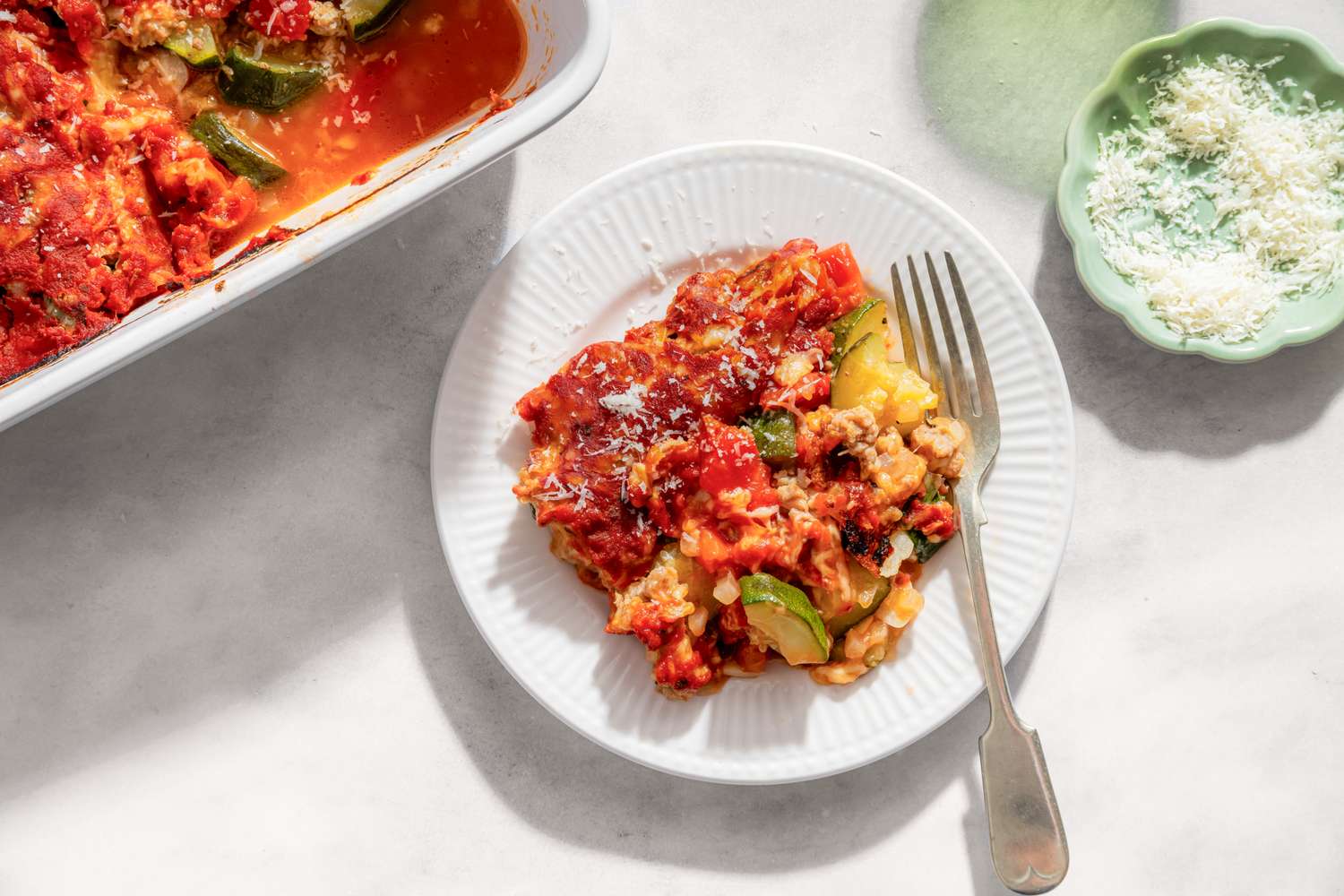 A plate of a zucchini pizza bake next to a casserole dish and a small dish of grated cheese