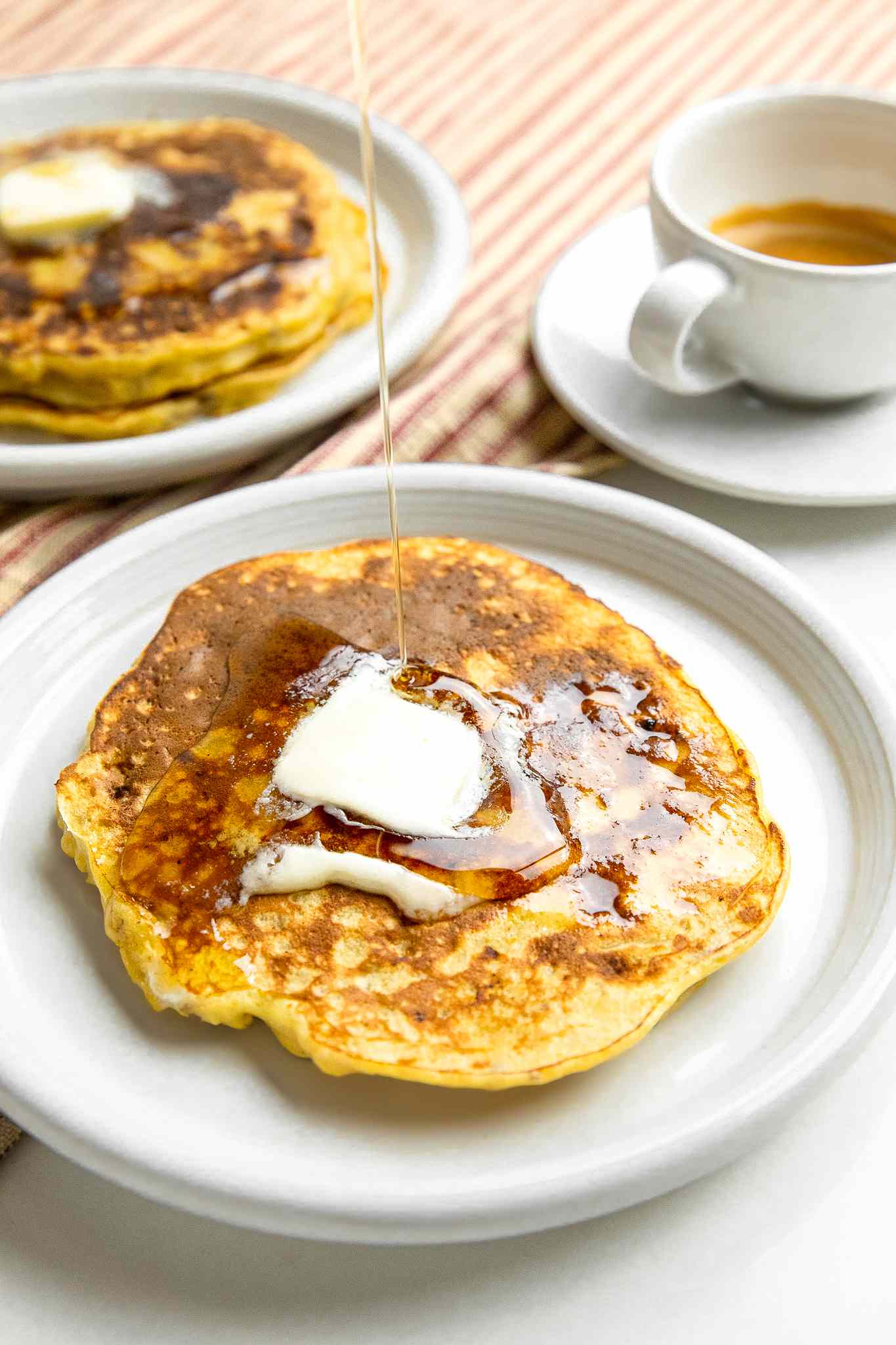 Syrup Drizzled onto a Plate with a Stack of 3-Ingredient Banana Pancakes with Butter, Surrounded by a Plate with More Pancakes and a Cup of Coffee