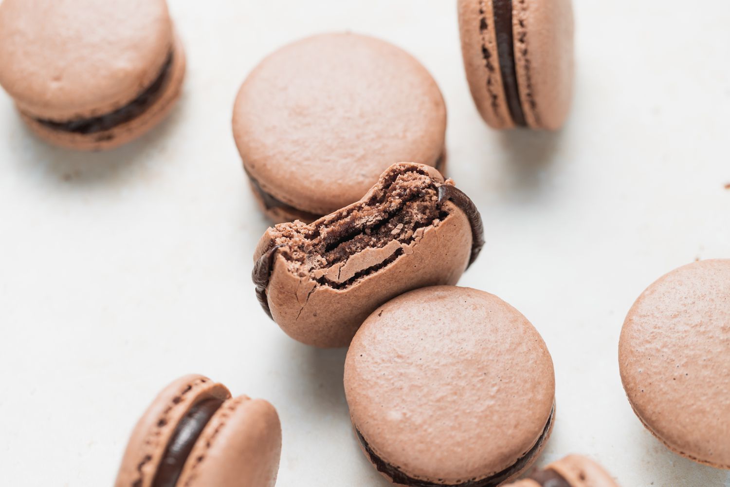 Overhead view of chocolate macarons scattered on a white background with the center one with a bite out of it.