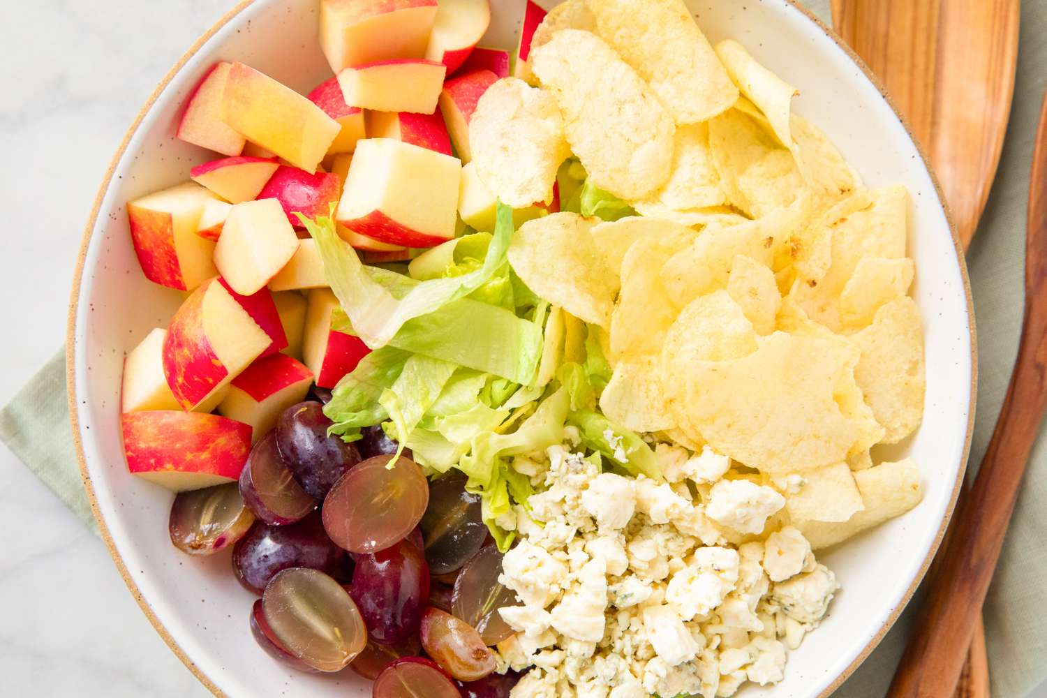 Bowl containing chopped apples, potato chips, lettuce, grapes, and cheese crumbles