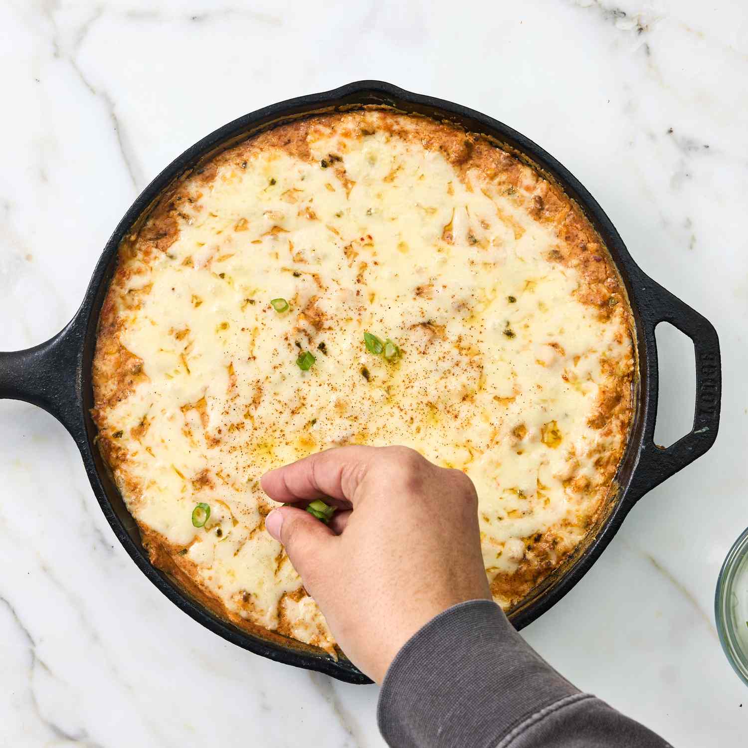 A hand garnishing a cheesy baked dish in a cast iron skillet with herbs