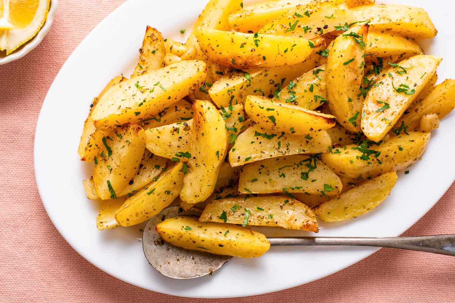 greek lemon potatoes topped with minced herbs on a plate next to a small bowl of lemon wedges