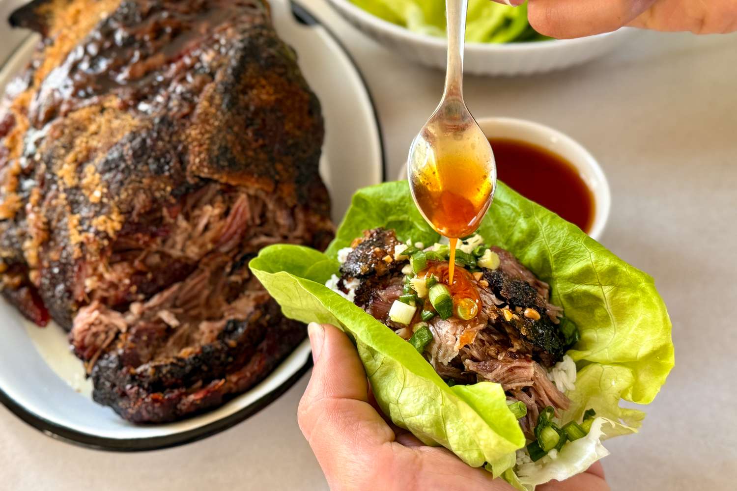 Shredded meat being prepared in a lettuce wrap sauce being drizzled from a spoon with a platter of meat and a bowl of sauce in the background
