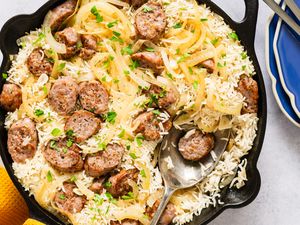 A skillet containing a sausage and rice bake with onions and fresh herbs a serving spoon resting inside