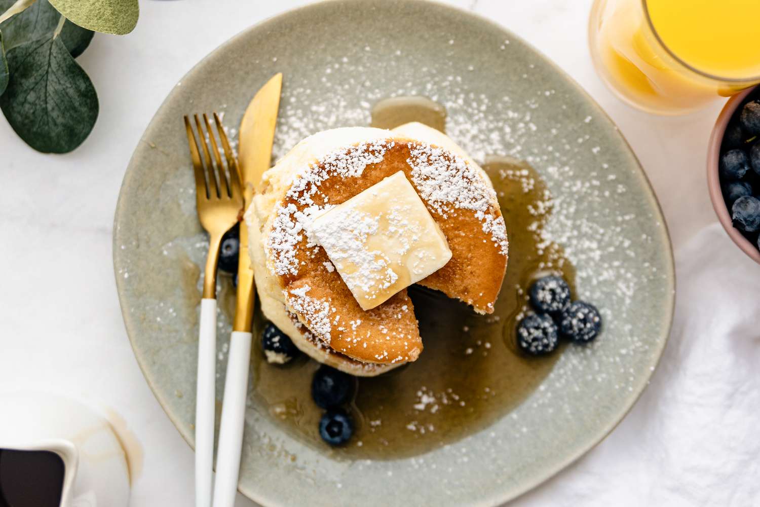 Aerial View of a Plate of Japanese Souffle Pancakes Topped with Butter, Maple Syrup, Powdered Sugar, and Blueberries with a Bite Sized Piece Cut Out
