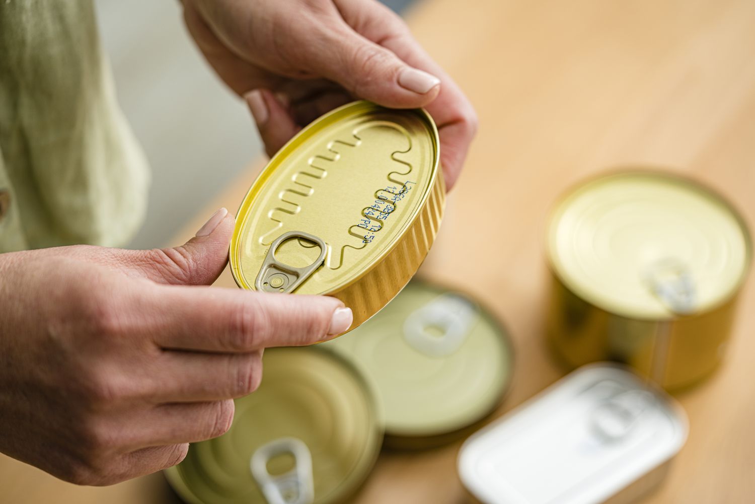 Person inspecting a sealed food can with other cans nearby on a wooden surface