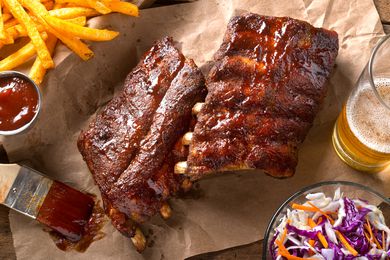 rack of glazed ribs on parchment paper at a table setting with a basket of fries, bowl of coleslaw, and a glass of beer 