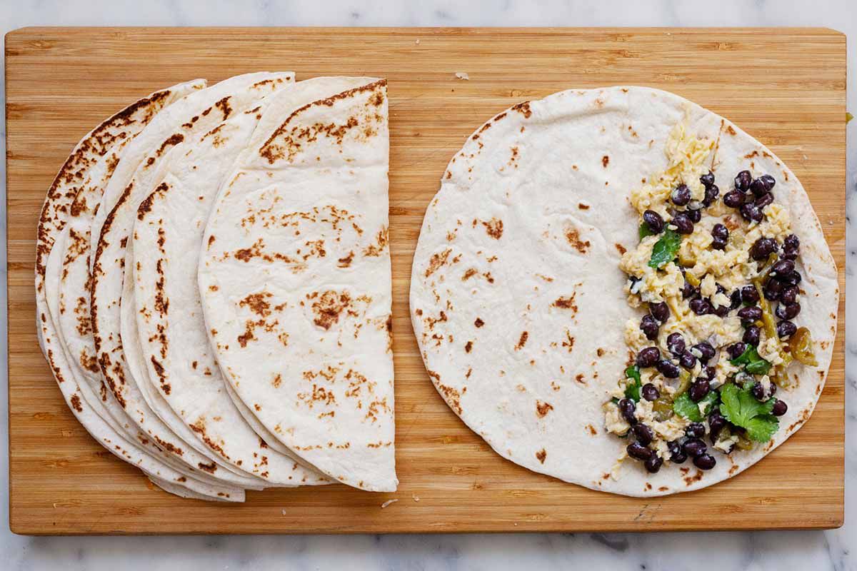 Four tortillas on a wooden cutting board for make-ahead breakfast quesadillas. One tortilla is open and has scrambled eggs, cilantro and bean filling on half. The other tortillas are folded in half with no filling inside.