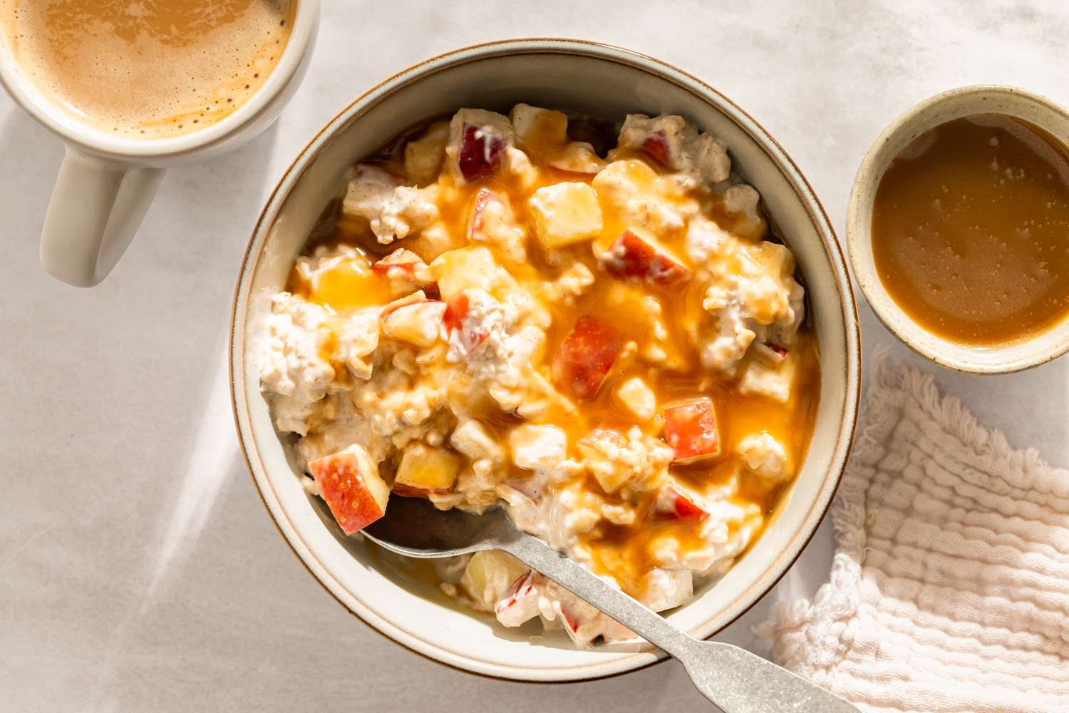 Bowl of overnight oats topped with chopped apples and caramel sauce with a spoon coffee and a small bowl nearby