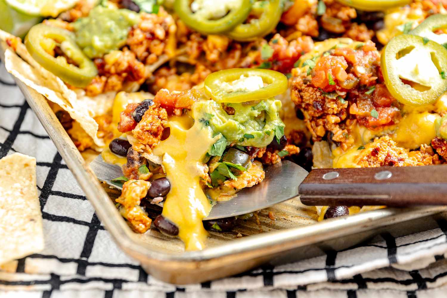 Baking Sheet of Vegan Nachos (Nachos Topped with Salsa, Queso, Pickled Jalpenos, and Two Lime Wedges) with Some Slightly Lifted on a Spatula