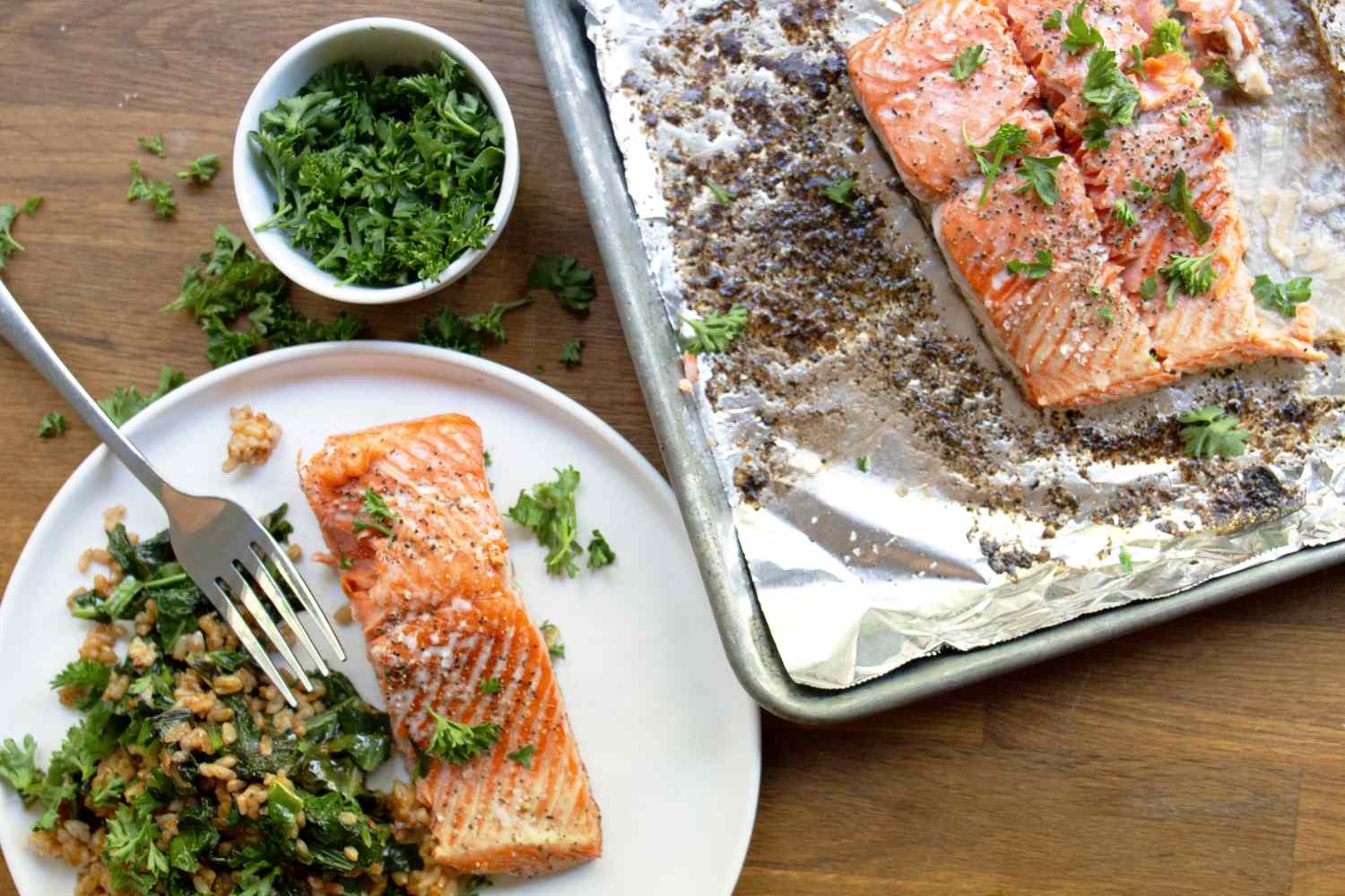 Cooked salmon on a plate with greens and a tray of prepared salmon nearby