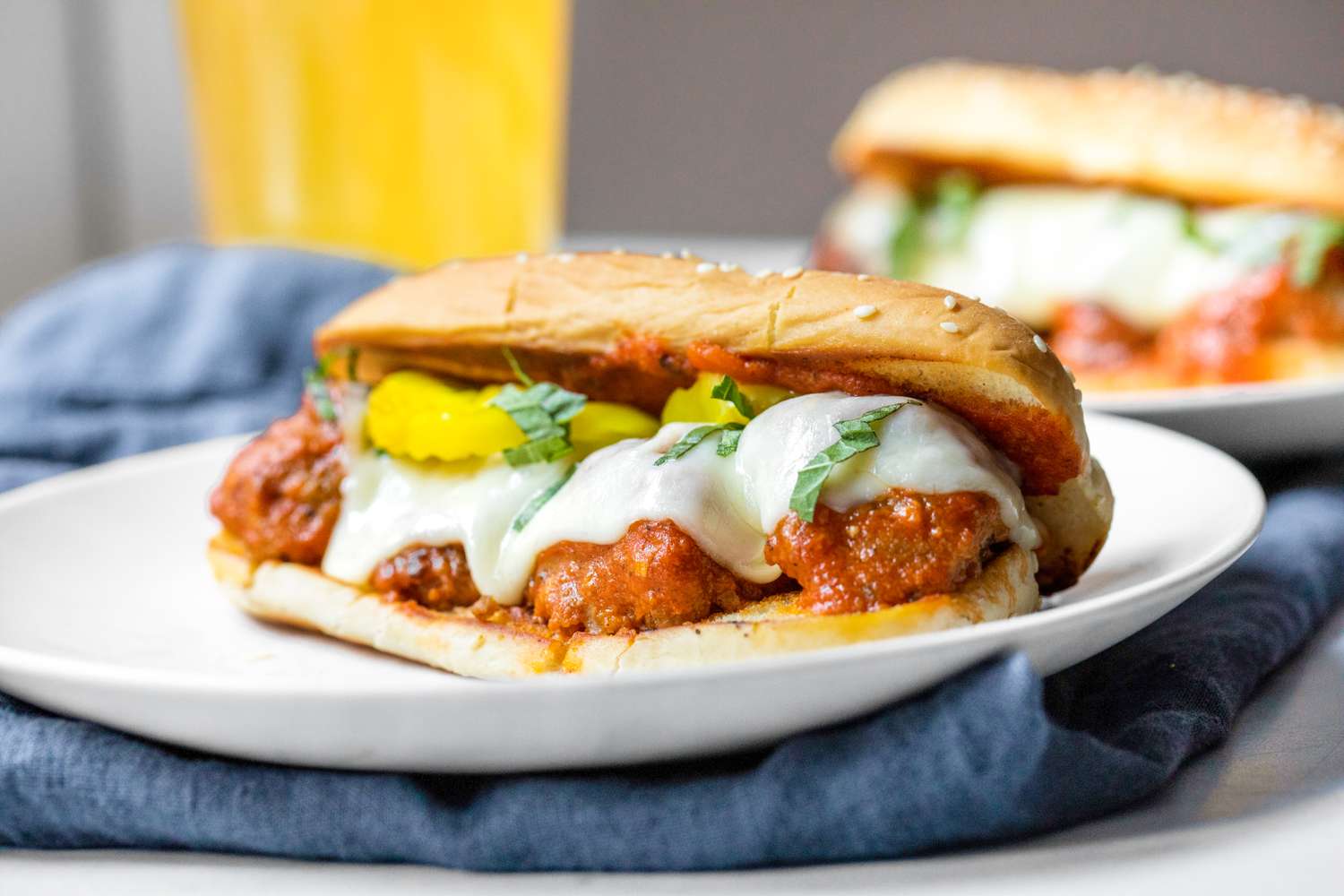 A Plate of Meatball Sub Topped with Banana Peppers and Basil, and in the Background, a Plate With Another Sub and a Glass of Beer