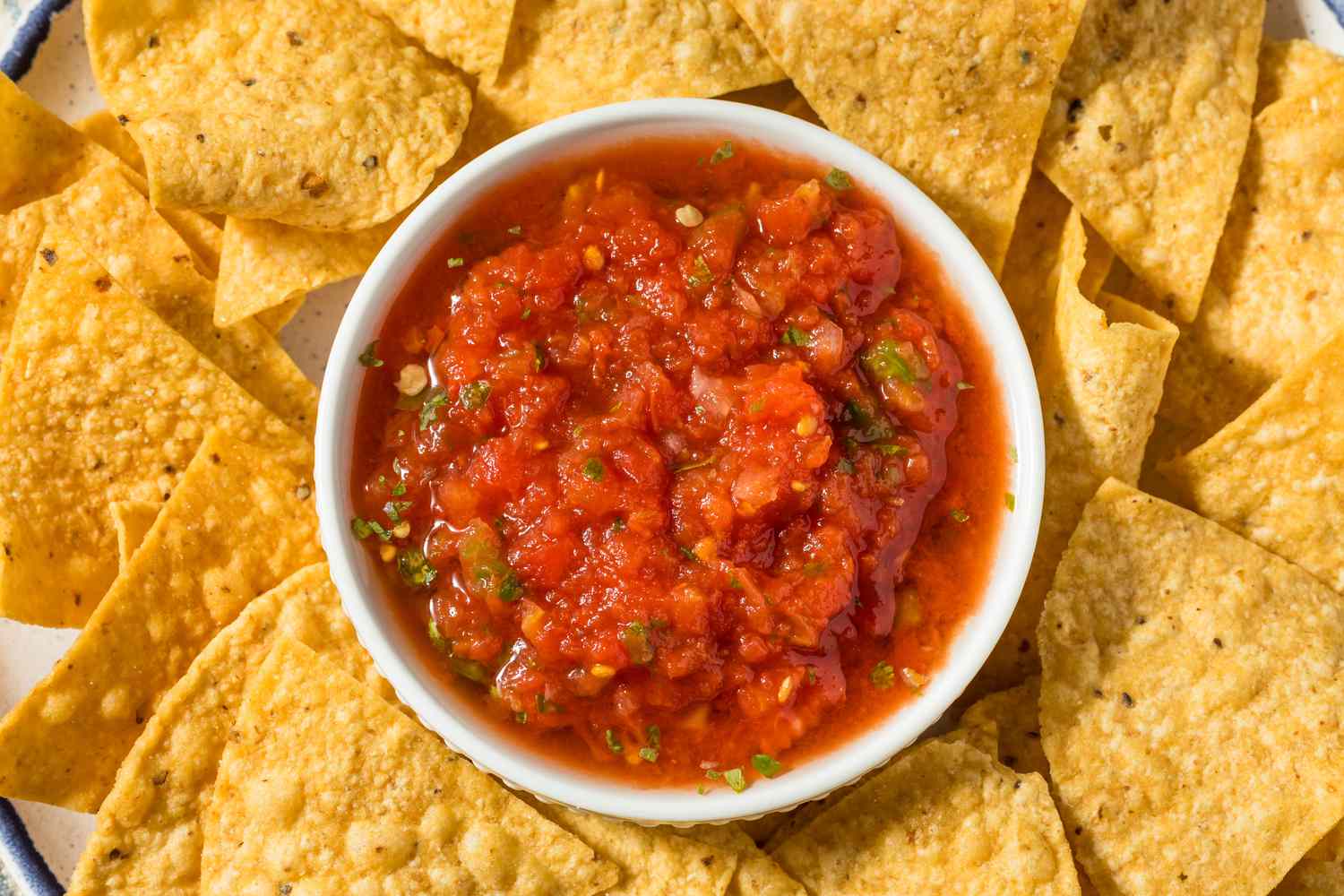 A bowl of red salsa in the middle of a plate of corn chips