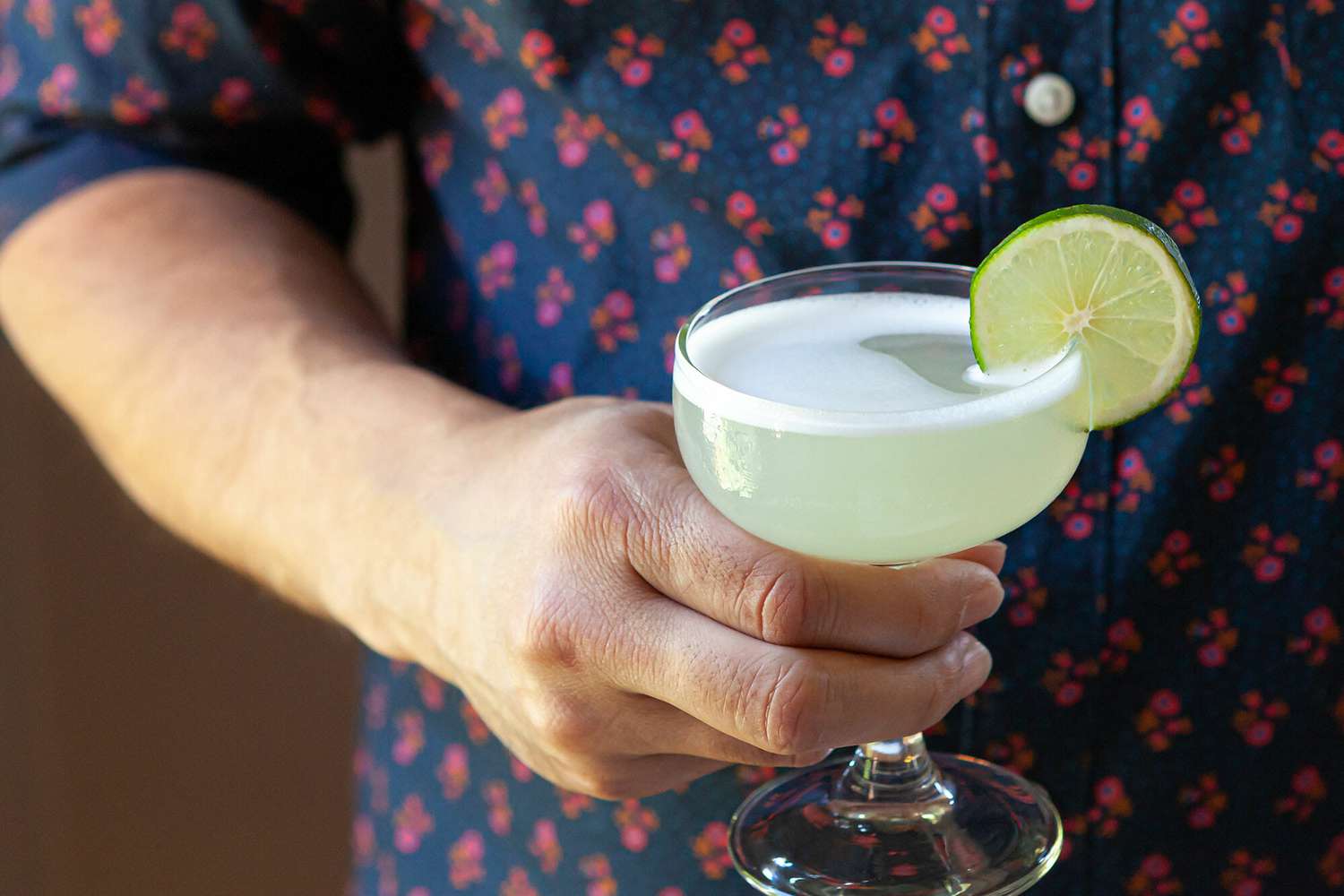 A person is holding a vodka gimlet in a chilled coupe with a lime wedge on the side.