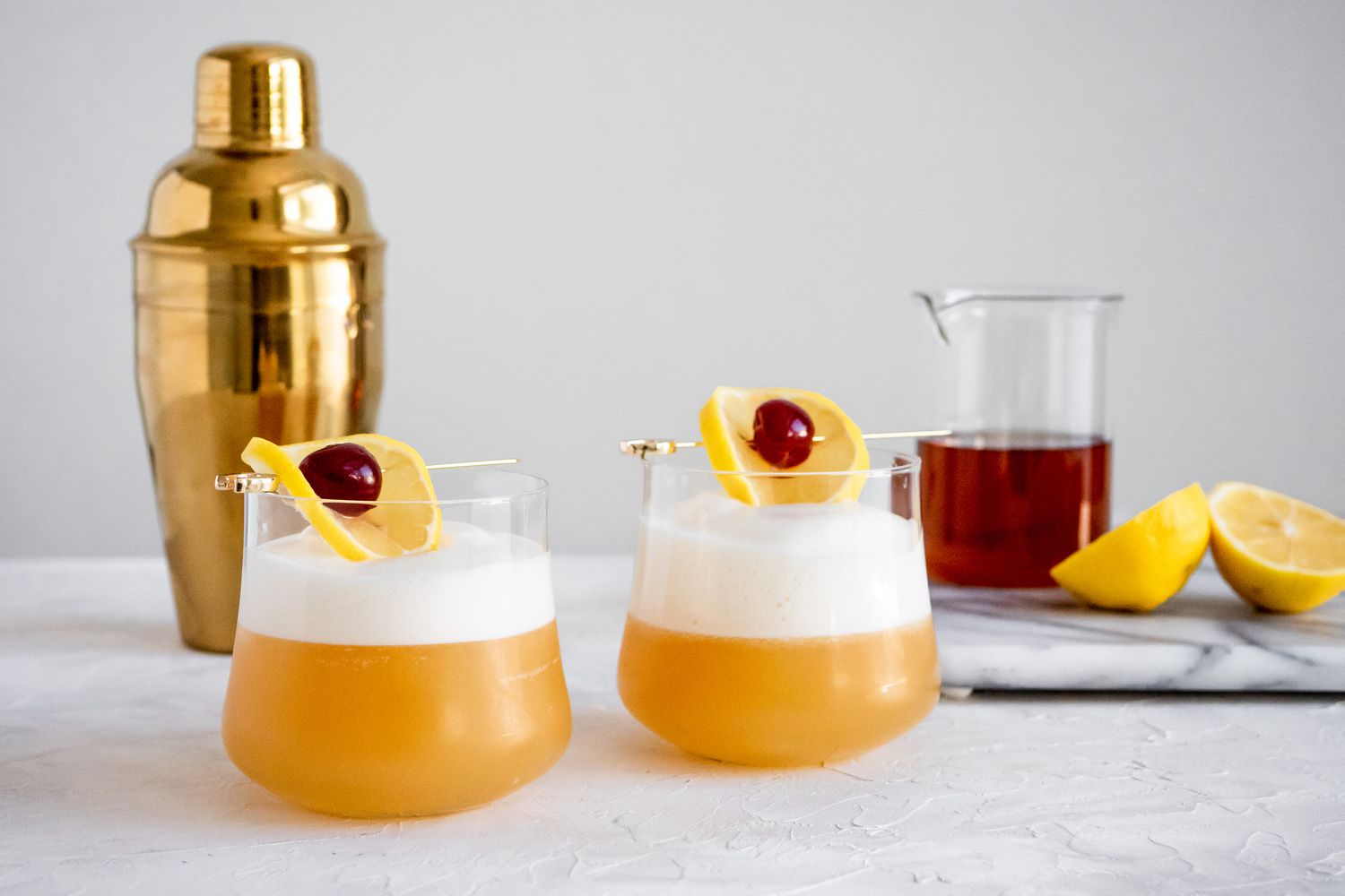 Two Glasses of Amaretto Sour with Ingredients and Cocktail Shaker in Background