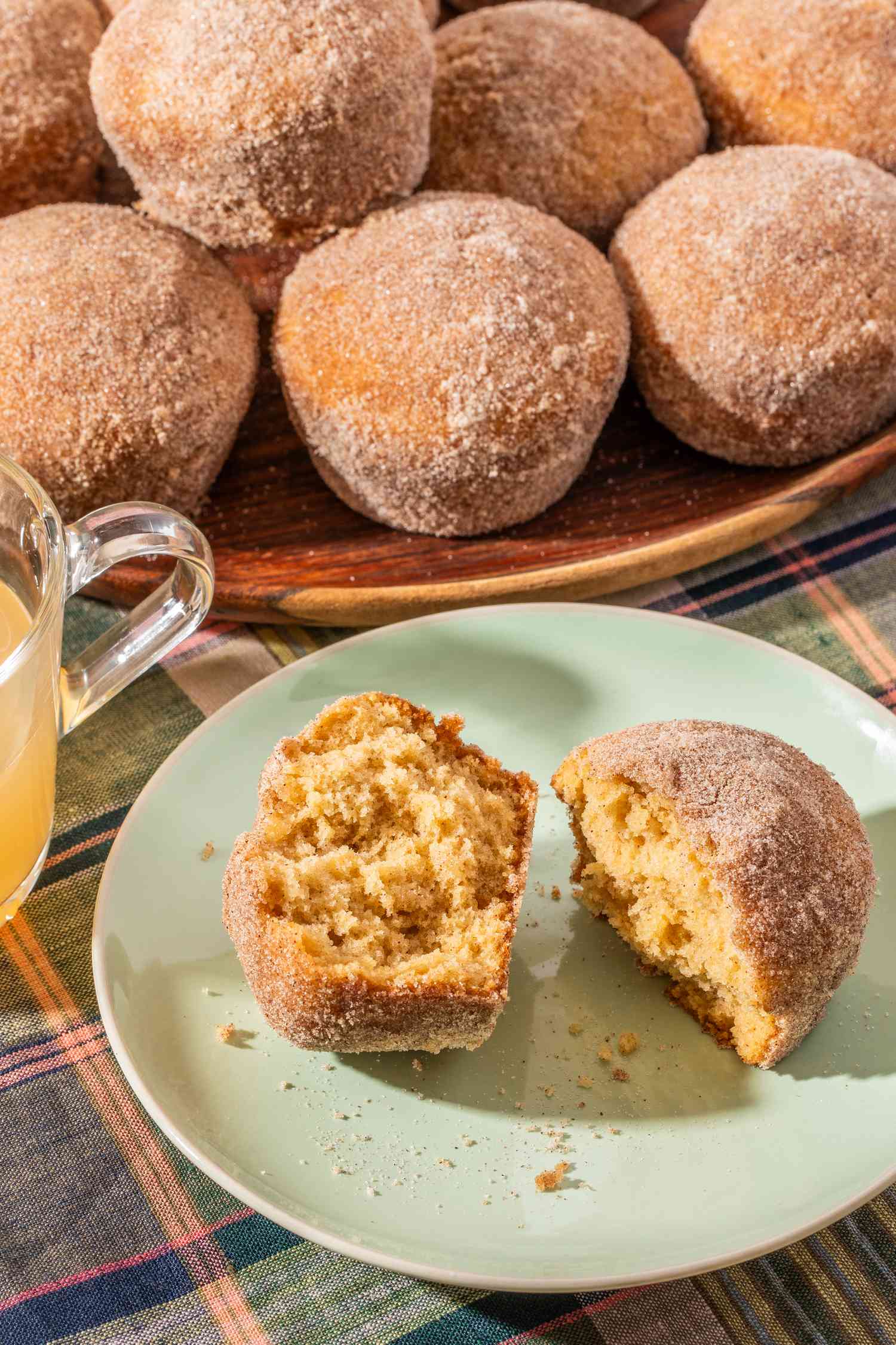 A halved apple cider donut muffin on a plate, and in the surroundings, a cup of apple cider and more muffins stacked on a plate
