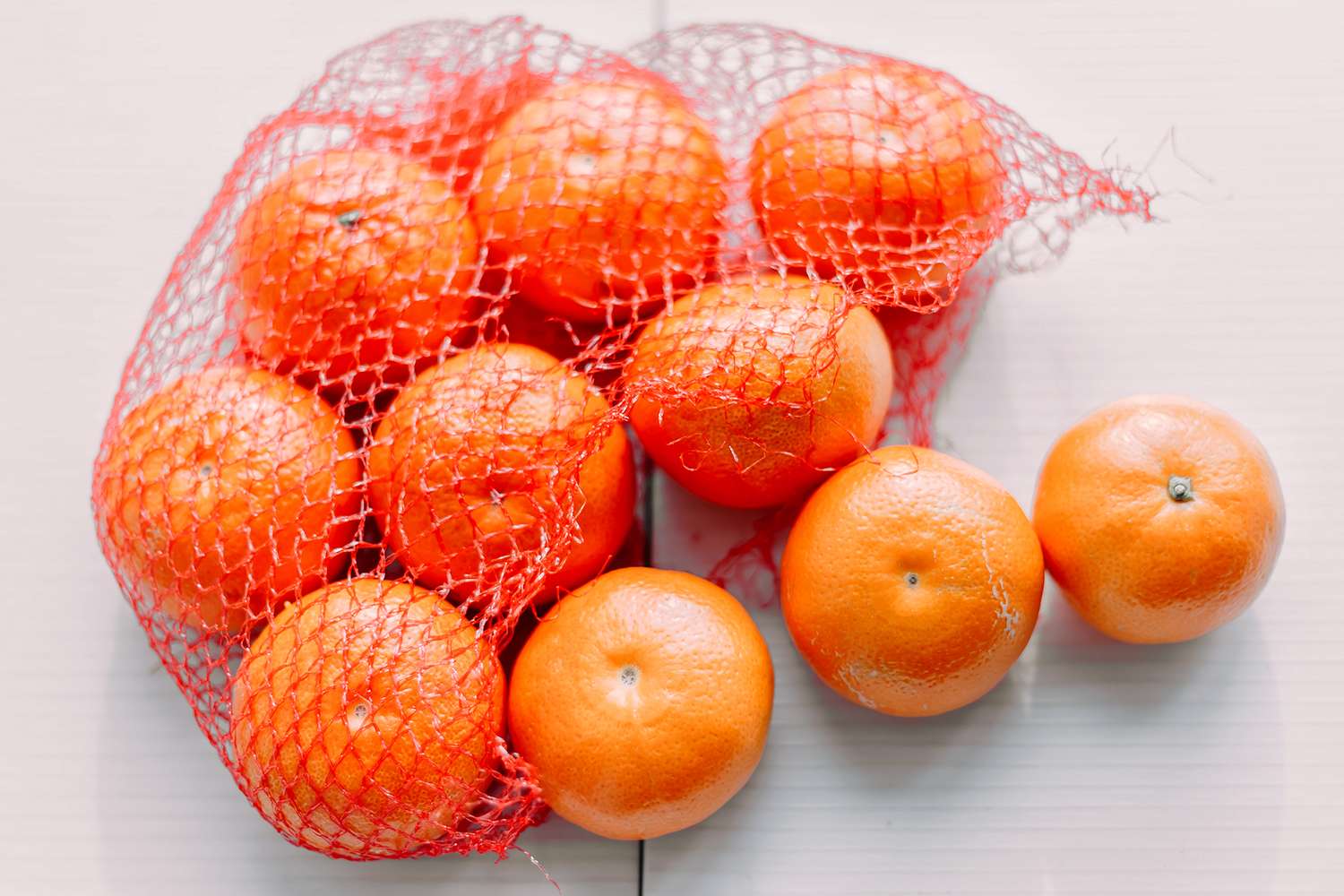 A group of oranges in a red mesh bag with three oranges outside the bag on a flat surface