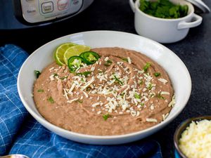 Pressure Cooker Refried Beans in a white bowl with lime wedges and sliced jalepenos nearby. The instant pot is in the background.