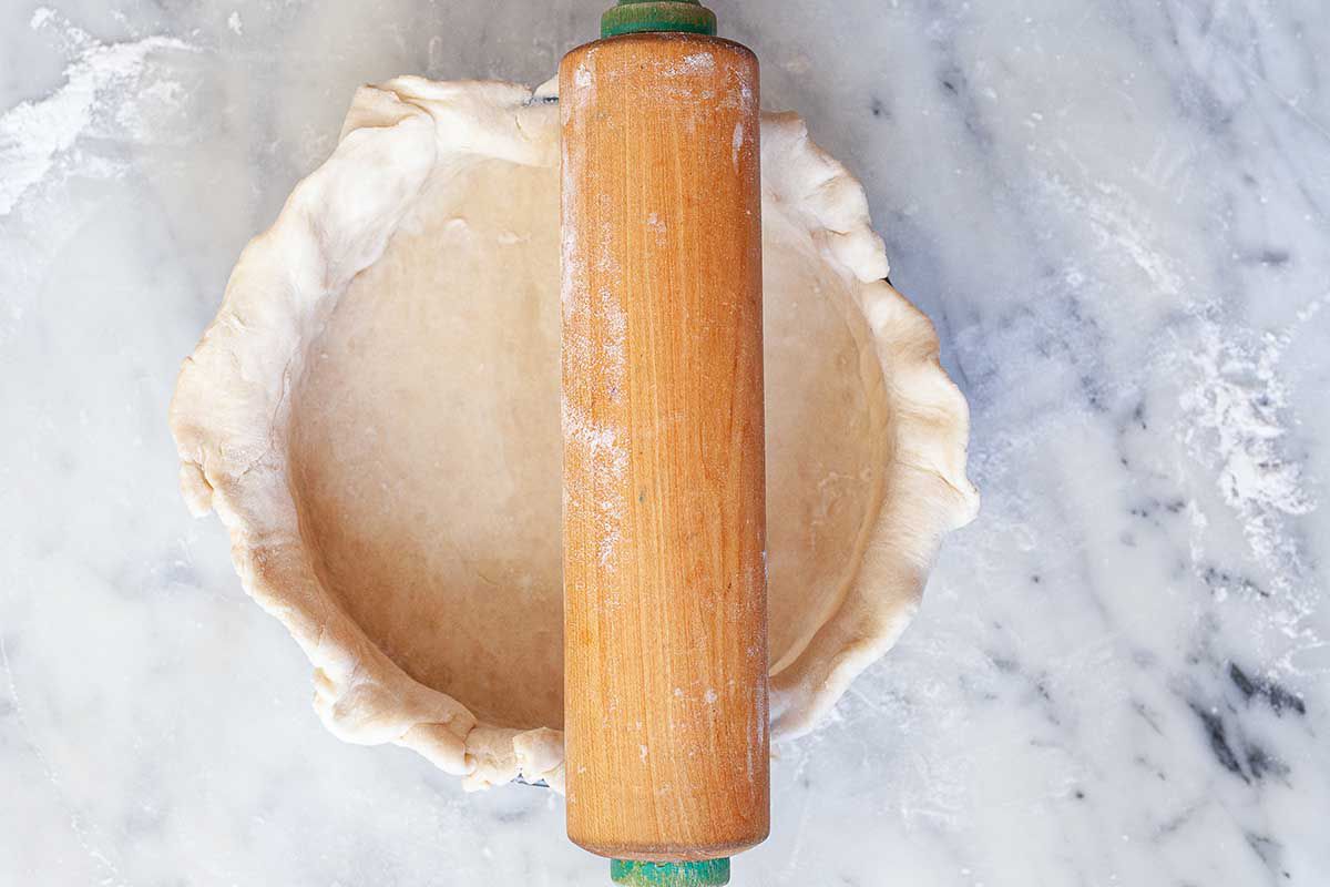A springform pan has an unbaked quiche crust inside. A wooden rolling pin is rolling over the top of the pan.