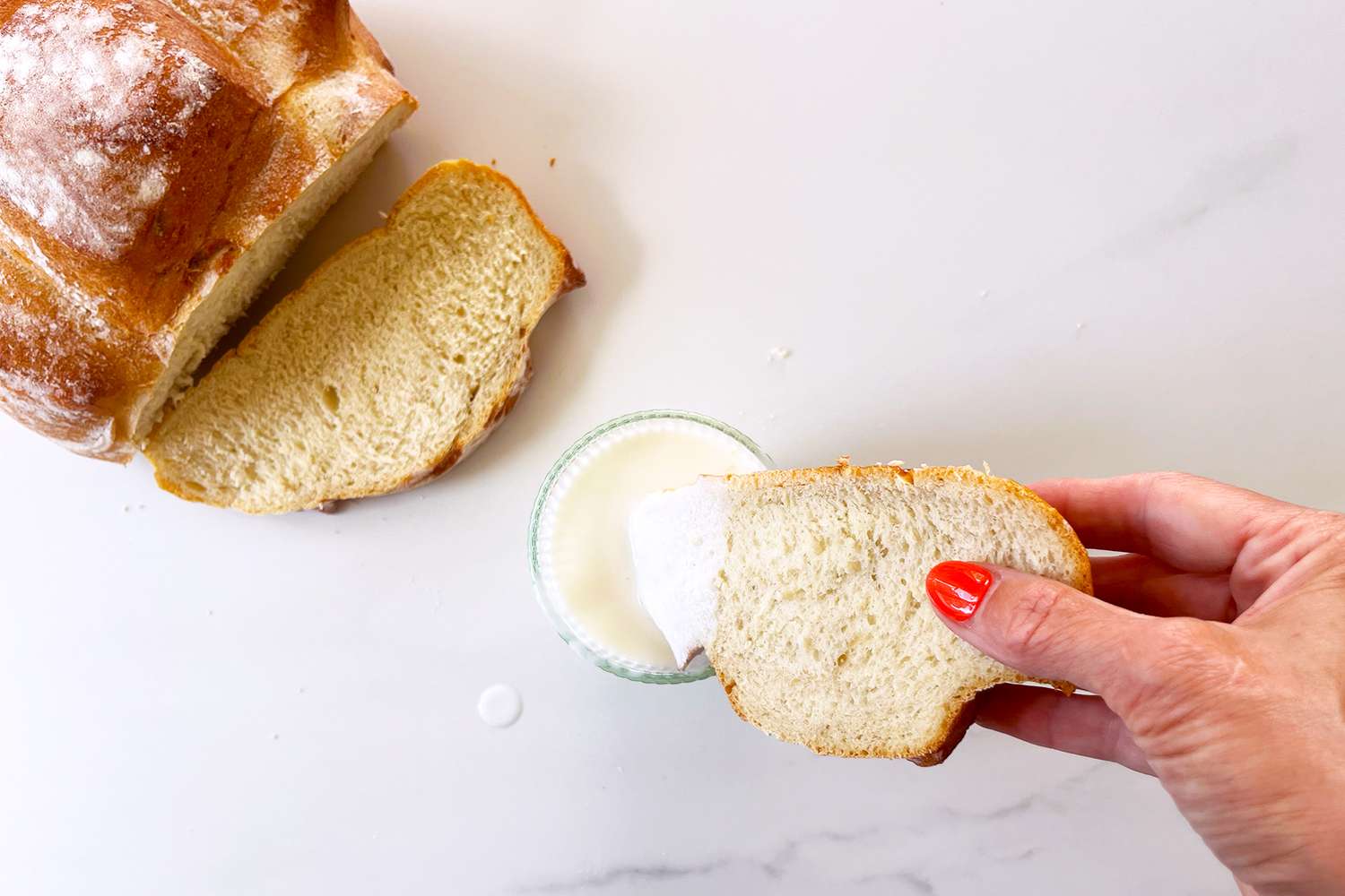 Hands dipping s slice of bread into a small dish of buttermilk, a loaf and a slice of bread in the background