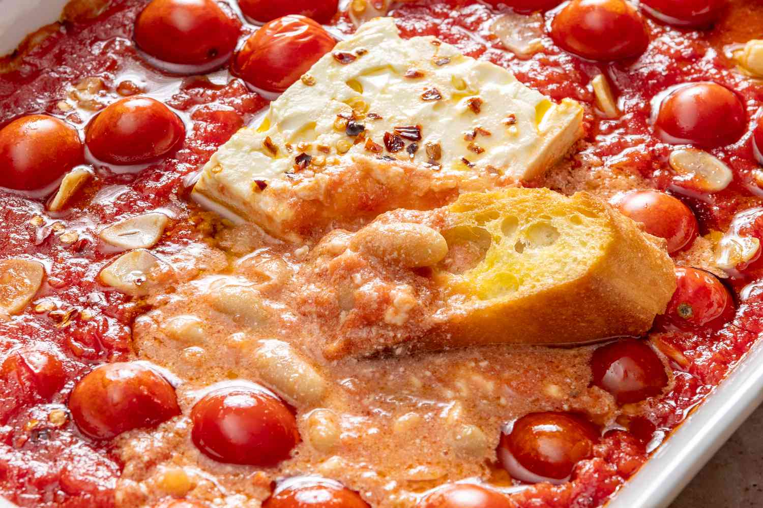 Close up view of a baking dish of garlic, tomatoes, cannellini beans and a block of feta covered in olive oil and red pepper flakes with a slice of french bread dipped into the mixture