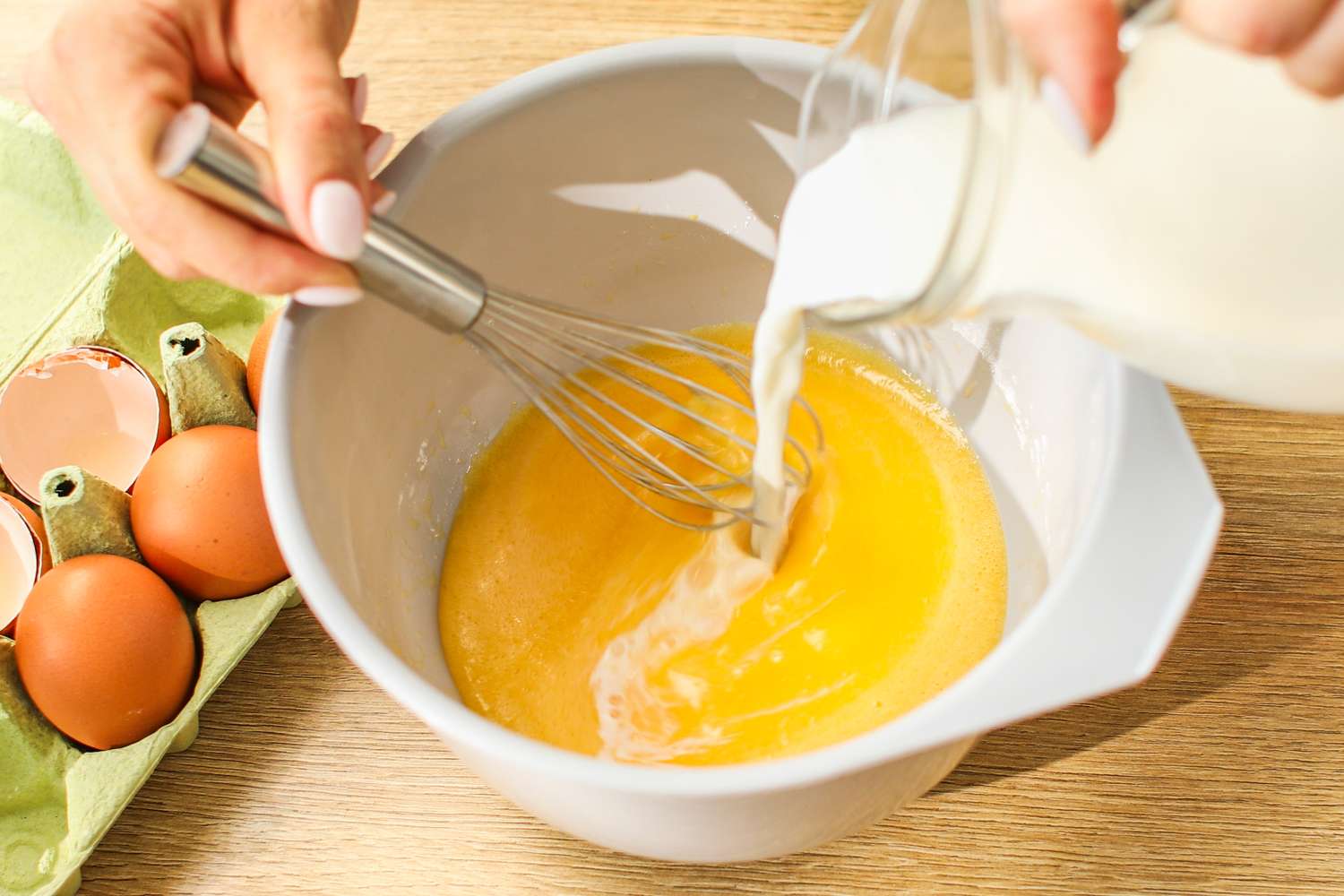 Preparing scrambled eggs with a whisk in a mixing bowl milk being poured into beaten eggs alongside a carton of eggs on the table