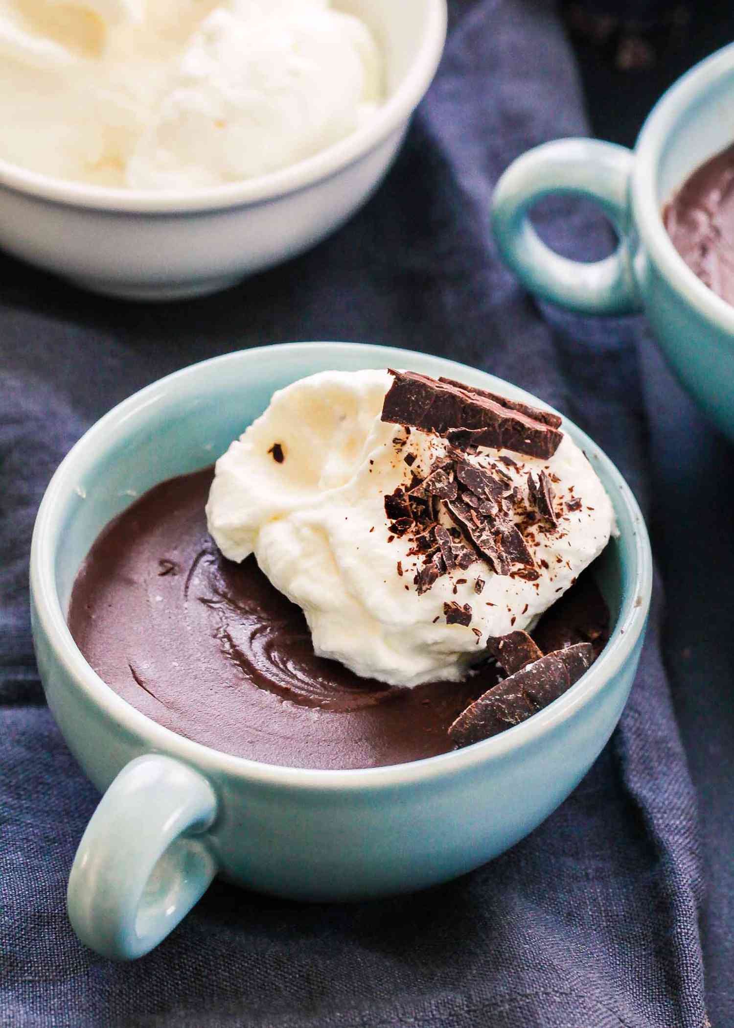 A serving of chocolate pudding in a ceramic cup topped with whipped cream and chocolate shavings